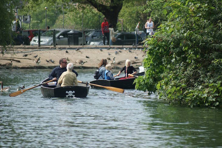 Two black row boats with people in them disappear around the side of an island in the boating lake in Finsbury Park