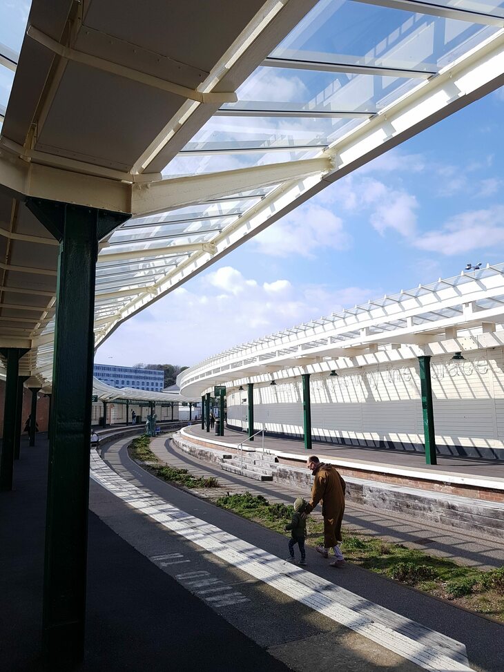 A curved former train platform stretching off into the distance is seen. The canopy is white with green plinths holding it up. The sky is bright blue with fluffy white clouds. A man and child can be seen walking along a walkway where trains would have run in the past, but is now a linear shingled garden and footpath.