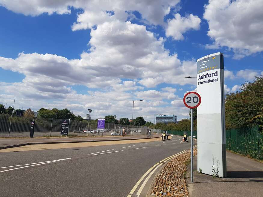 A grey upright sign with the words Ashford international, and the Eurostar logo is seen on the right hand side of the image. A 20mph sign is in front of this. A road runs off into the distance with a largely empty carpark to the left. The sky is beautifully blue with fluffy white clouds. 