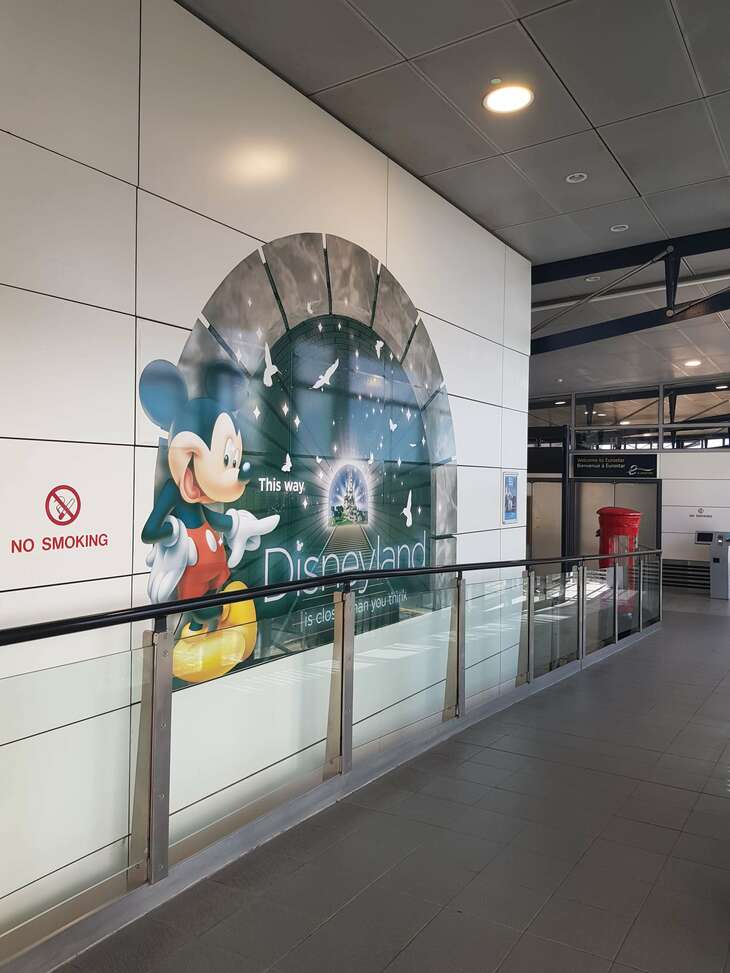 A Disneyland sign is seen on the wall of a station. It shows Mickey mouse pointing to the right of the image with the words "Disneyland. Closer than you think" underneath