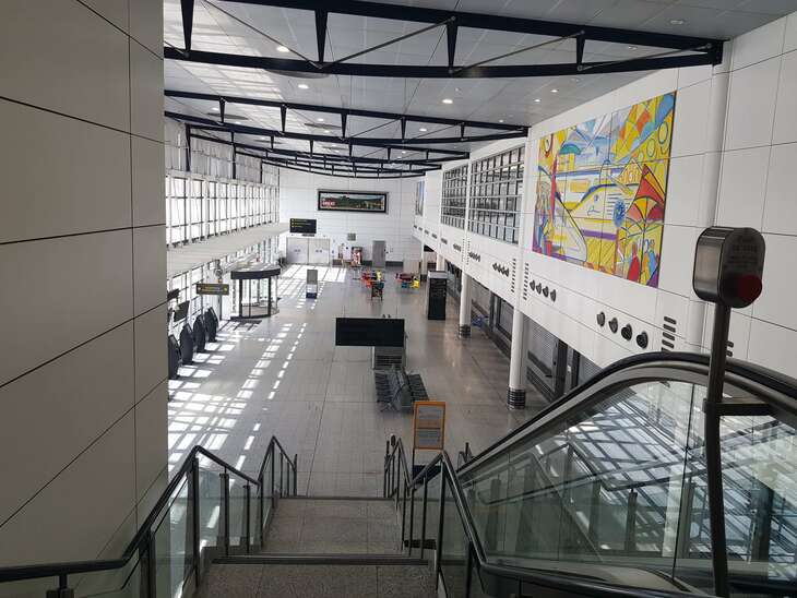 The international entrance to Ashford International station is photographed from the 2nd floor of the building. A large cavernous terminal can be seen with no people visible. The handrail for the escalators is visible in the foreground, and a disused cafe in the background. 
