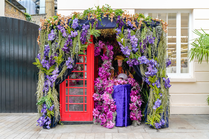 A red phone box inside a purple gazebo, with a curtain of purple and pink flowers draped in front of it