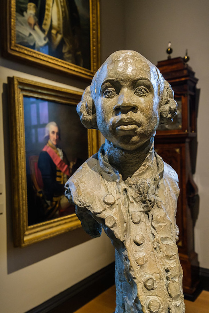 A bust of Equiano in a museum with a portrait of a bewigged naval gentleman in the background
