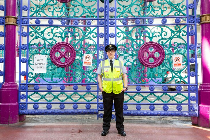 A person in high vis poses in front of ornate blue, purple and teal gates