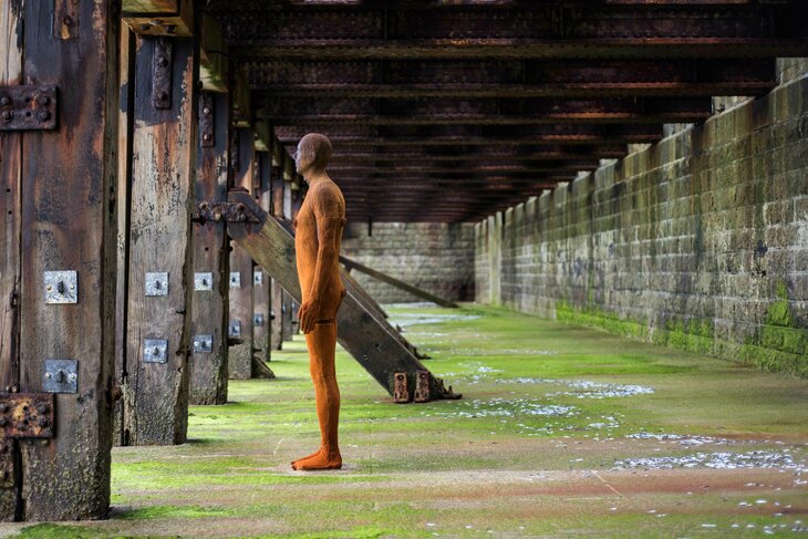 A rusted metal figure is staring out into the distance. It appears to be underneath a wooden walkway and standing on a weathered, green concrete standing. 
