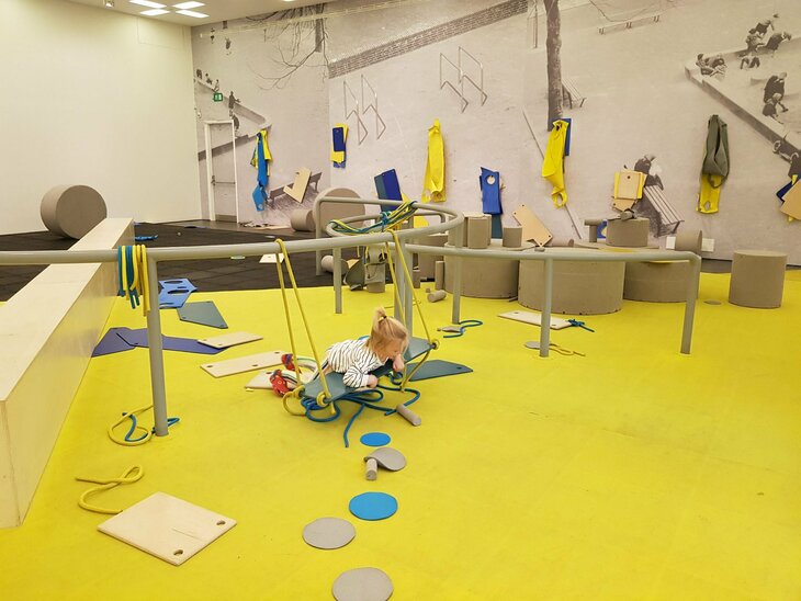 A child is playing on a homemade swing in the middle of an interactive art exhibition. The exhibition consists of rails, ropes, large foam circles, wooden planks and fabric bands, all placed around the room for viewers to interact and build with. 