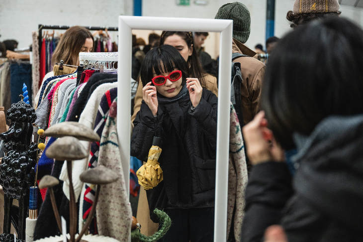 A woman trying on a red pair of glasses in a mirror, in front of rails of clothing.