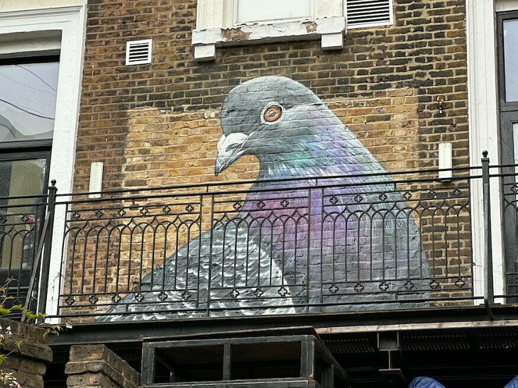 The head and upper body of a pigeon, street-arted onto a brown brick wall with a metal balcony in front