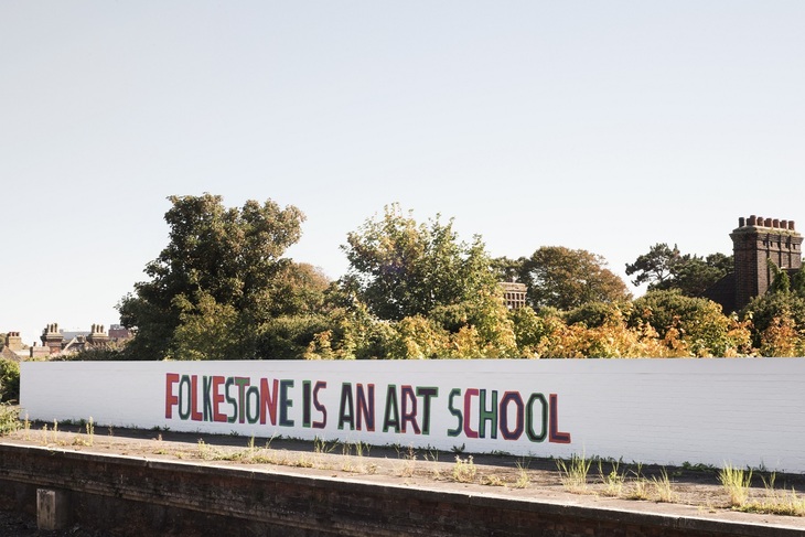 It is a sunny day. "FOLKESTONE IS AN ART SCHOOL" is painted in multiple colours on a white wall next to a slightly overgrown train platform. 