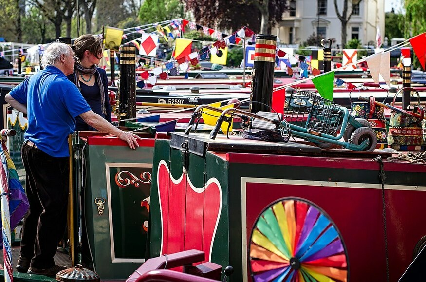 Canalway Cavalcade: a couple stand on their narrowboat in Little Venice, surrounded by other boats, all decked out with colourful flags and bunting.