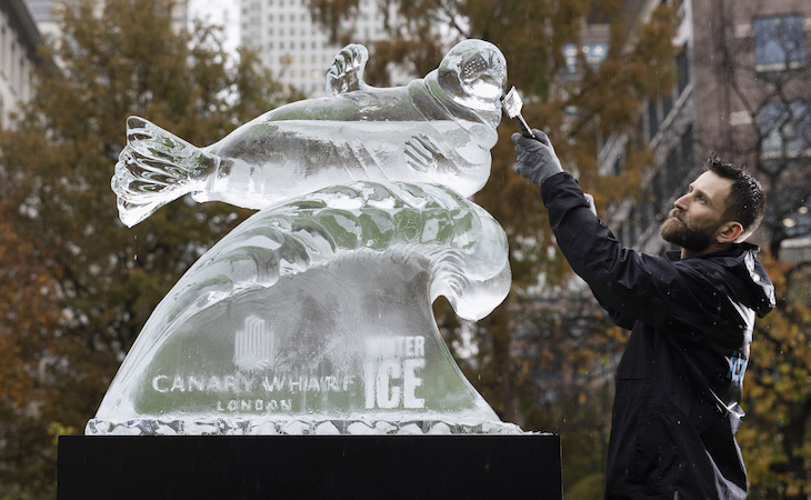 A man sculpting a seal from ice, with the One Canada Square tower in Canary Wharf in the background