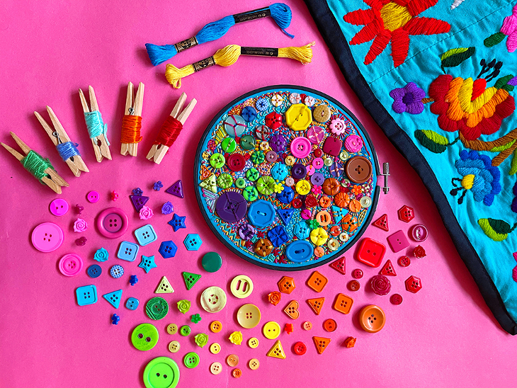 An embroidery hoop covered in colourful buttons on a table, surrounded by more buttons, thread, and other craft materials
