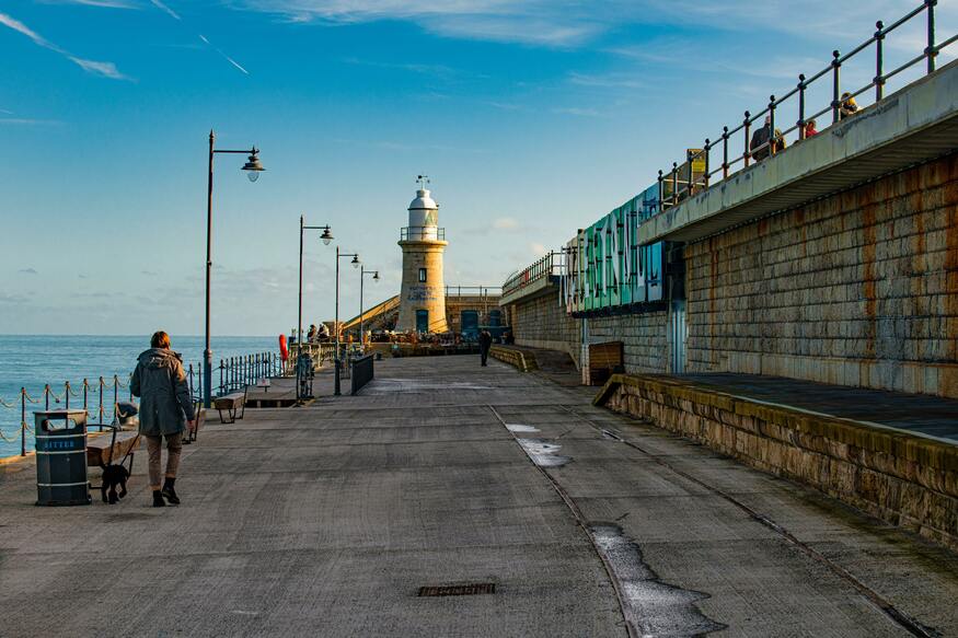 It is a sunny winters day. The photo is of an old restored harbour arm. At the end of the phot is a small granite lighthouse, now a champagne bar. To the right of the image is a brick wall, and to the left is the english channel. People are seen walking along the length of the walkway of the harbour arm