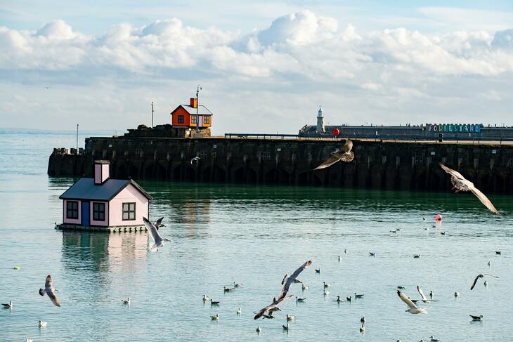 Seagulls are swooping down over an expanse of water. There are 2 cartoon style houses seen. One is floating in the water and is pale pink in colour. The second is perched on a plinth on dry land in the background. The harbour arm and lighthouse of folkestone can be seen in the background. 