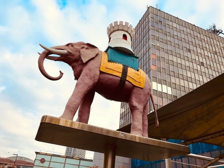 A pinkish elephant with a castle on its back, in front of a 1960s tower block