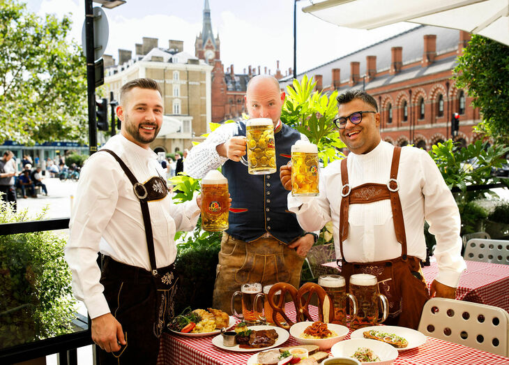 Three men in lederhosen drinking steins of beer with a table of German food in front of them