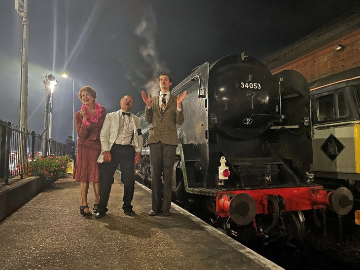 Actors dressed as Basil, Sybil and Manuel from Fawlty Towers standing on a train platform in front of a steam train