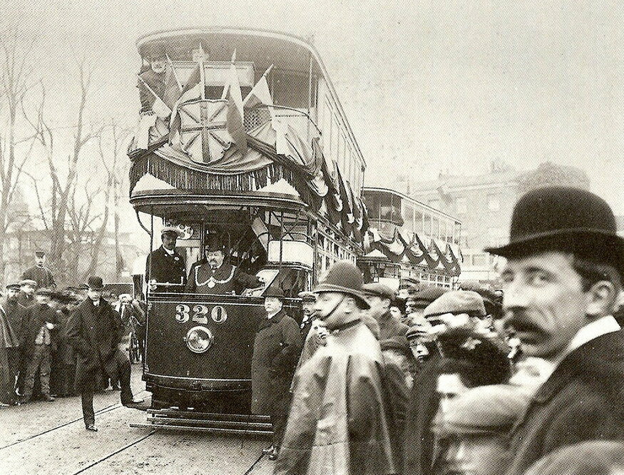 A festooned tram surrounded by onlookers including a chap in a bowler hat close up, looking at the camera