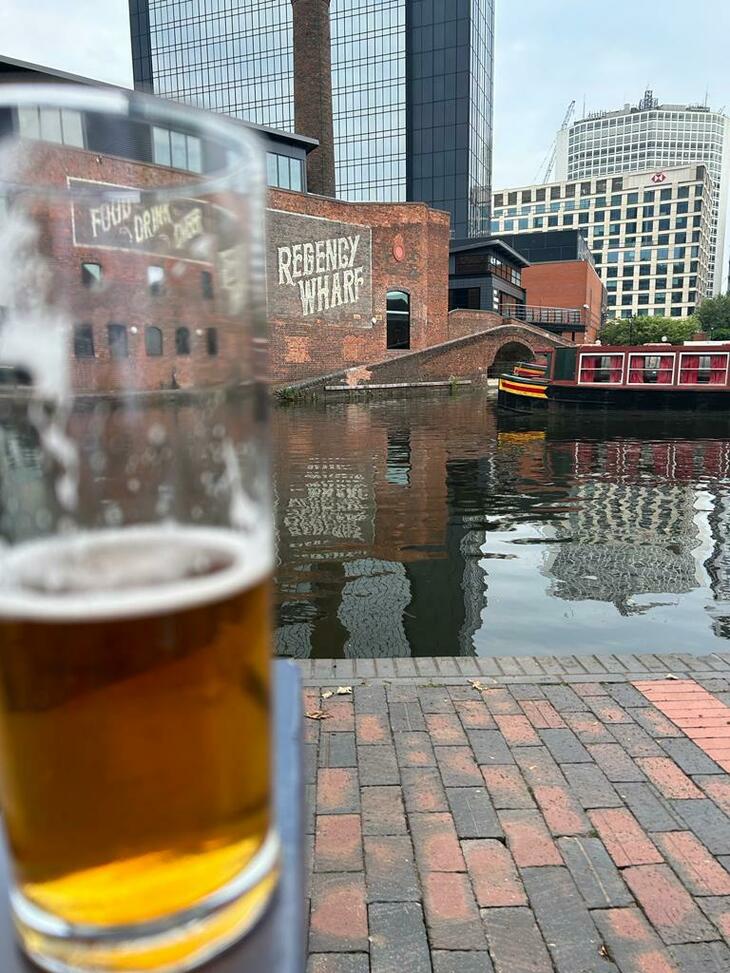 Things to do in Birmingham: A pint of beer in front of a canal with a redbrick warehouse and narrowboat sailing past