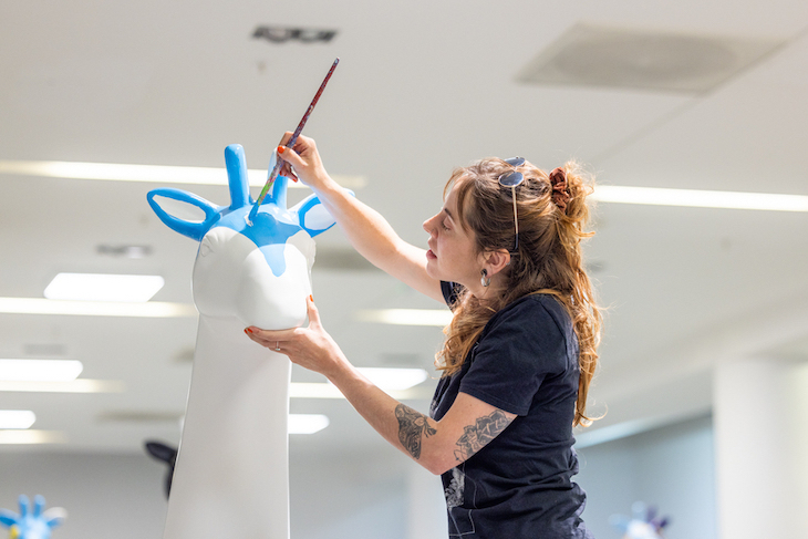 A lady paints the head of a white model giraffe with blue paint.