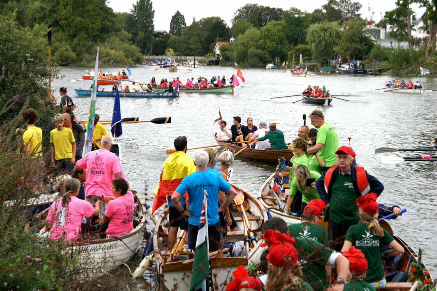 An assortment of small boats on the Thames, loaded with people wearing various brightly coloured clothes