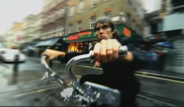 Ian Brown riding a curve-handled bike across a Soho street in the rain