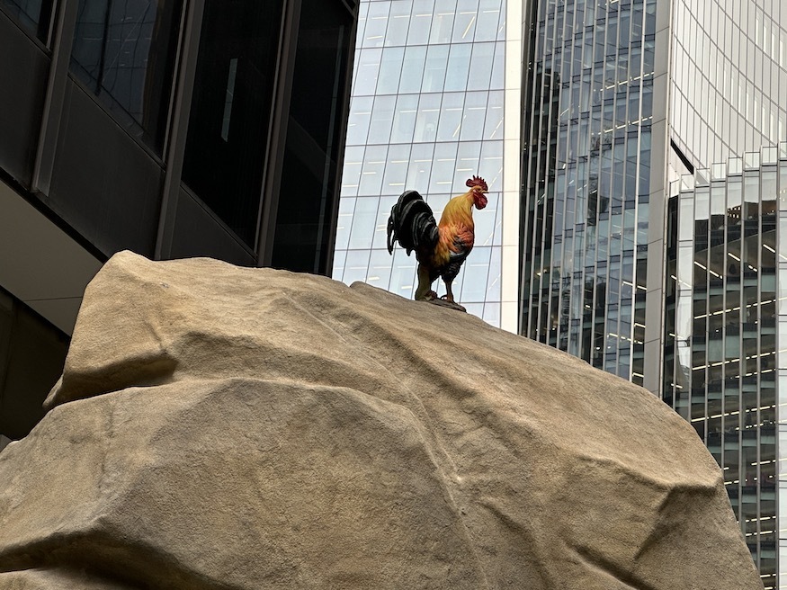 A sculpture of a cockerel perching on a fake rock with skyscrapers behind