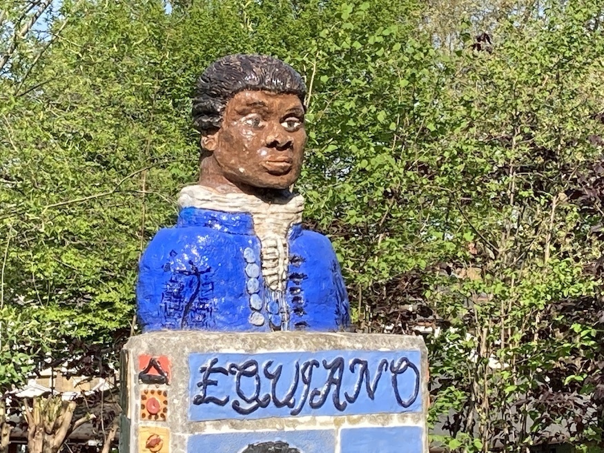 A blue-shirted bust of Oulaudah Equiano on a plinth. Greenery in background