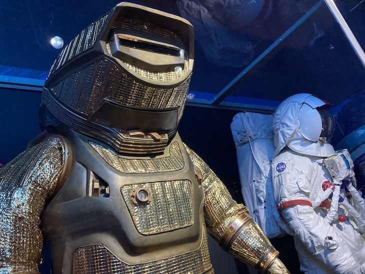 Two spacesuits. The one on the left is heavily shielded and golden. That on the right is a white Apollo moon suit