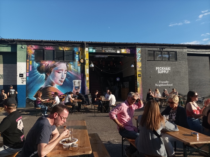 Brewery taprooms in London: People eat and drink in a sunny courtyard, backdropped with the Kanpai warehouse, painted with a large geisha-type figure