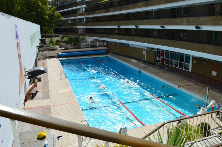 A bird's eye view of people swimming in the outdoor pool at Oasis Sports Centre, which is overlooked by a block of flats