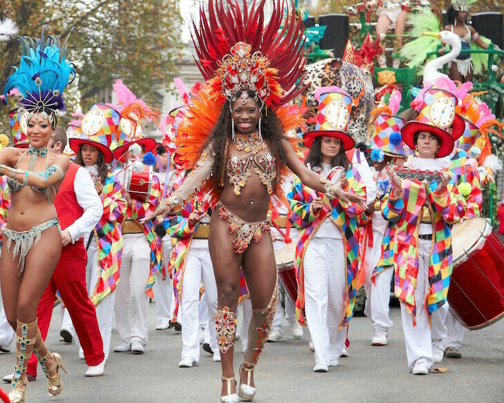 A woman dressed in a Carnival-style costume with a red headdress in the Lord Mayor's Show procession, with other performers in different costumes visible behind her