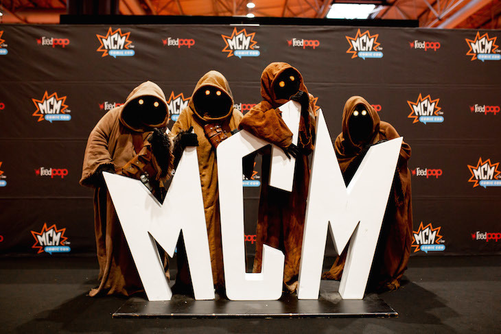 Four people in hooded cloak costumes pose next to giant MCM letters