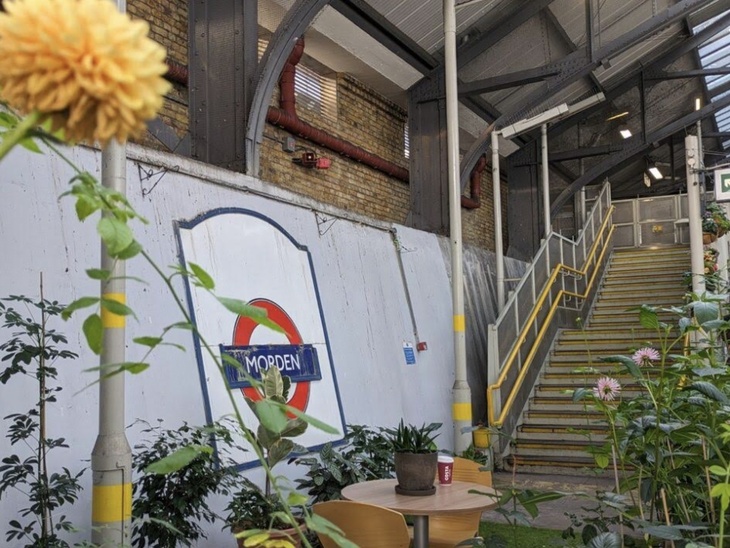 A garden in the foreground, with a Morden roundel and platform steps behind