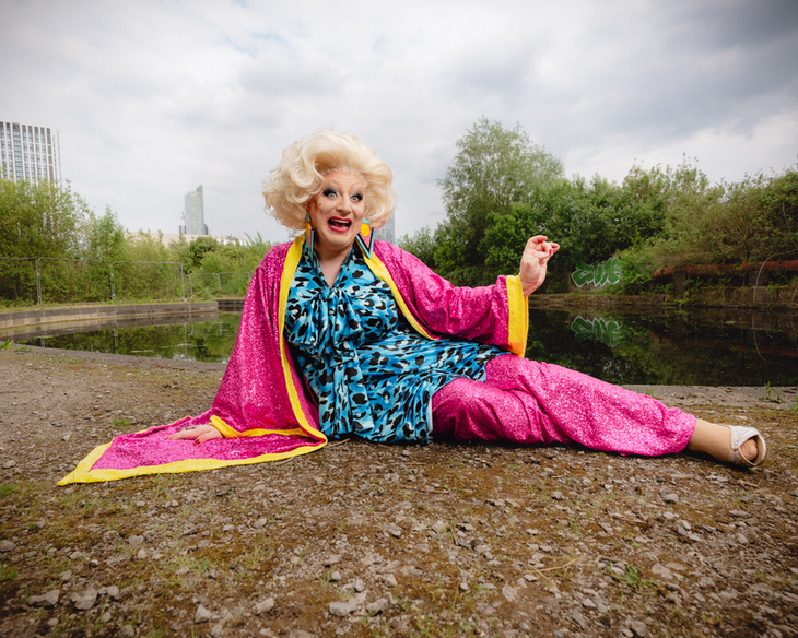 A drag queen sprawled in front of a lake, wearing a chocking pick gown