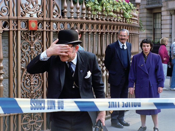 A man in front of police tape, wearing a bowler hat, covers his eyes