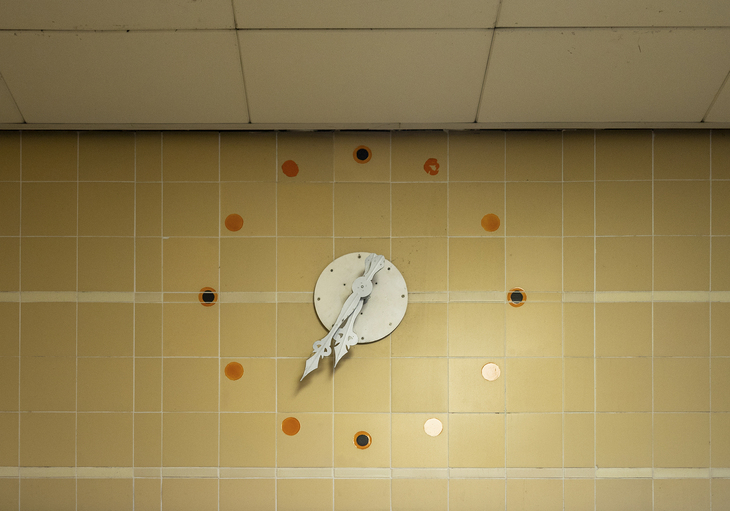 A clock on a beige tiled wall - with little else but hand and pips stuck on the tiles
