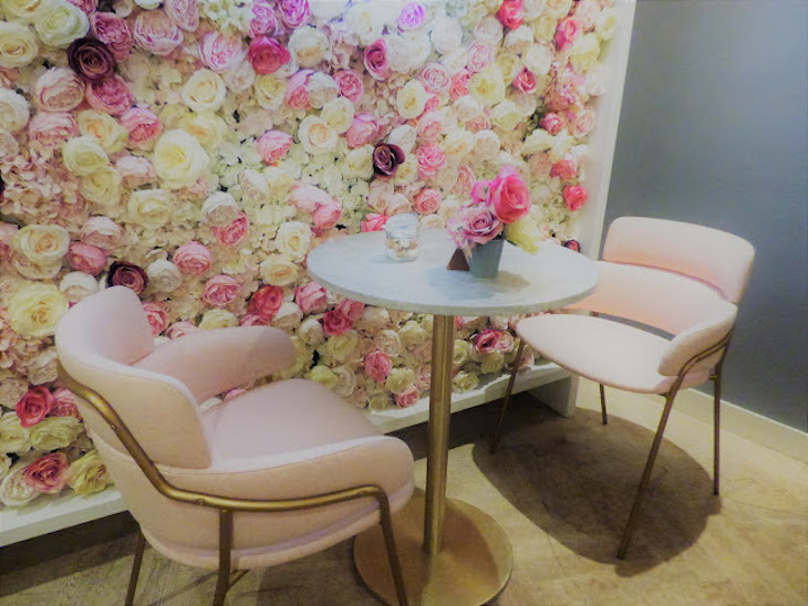 Two chairs and a small table alongside a flower wall consisting of white and pale pink roses, inside a cafe.