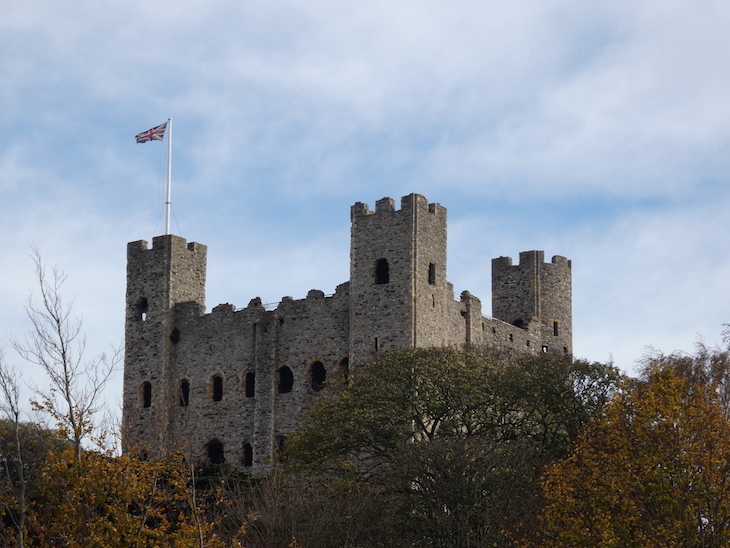 The exterior of Rochester Castle, a square stone building with square towers at each corner and a Union Jack flag flying from the roof