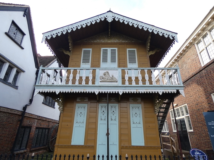 A two-storey wooden chalet painted a ginger-brown colour, with window shutters and other trimmings painted a light blue