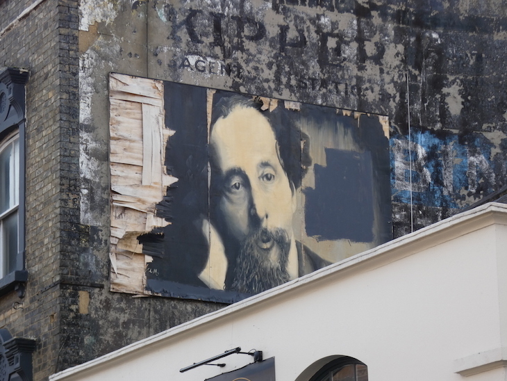 A mural of Charles Dickens looking over Rochester High Street, plastered onto a building over the top of advertising ghost signs