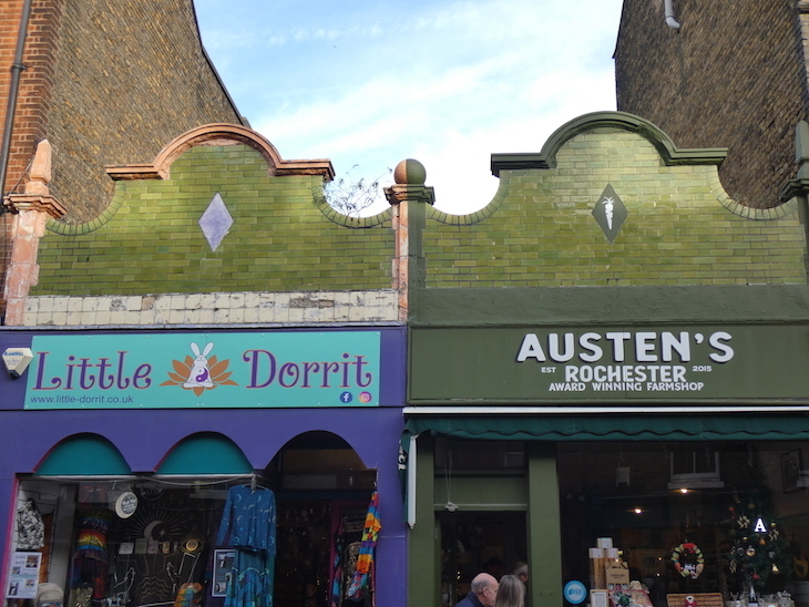 Exterior of Little Dorrit's gift shop and Austen's greengrocers next door to each other, with green tiled arches above their shop signs