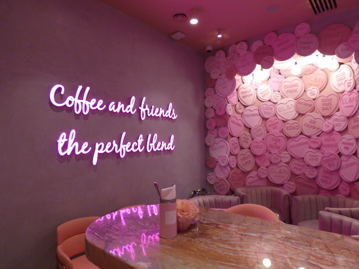 A wooden table in a cafe. One wall behind it is pink, with pink neon writing saying 'Coffee and friends the perfect blend'. The other wall is covered in oversized pink Love Hearts sweets.