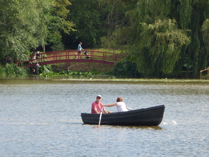 Two people in a row boat in the middle of the Hever Castle lake, with a man with a pushchair walking over an arched footbridge in the background