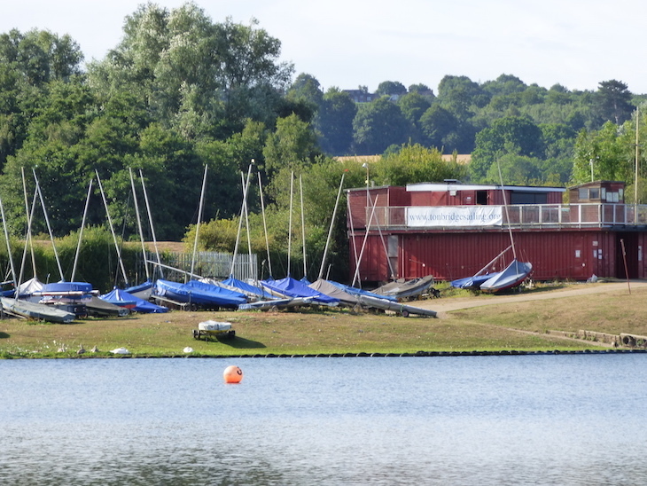 Looking across a lake to a group of sailing boats moored on a grassy area, next to a wooden boating shed.