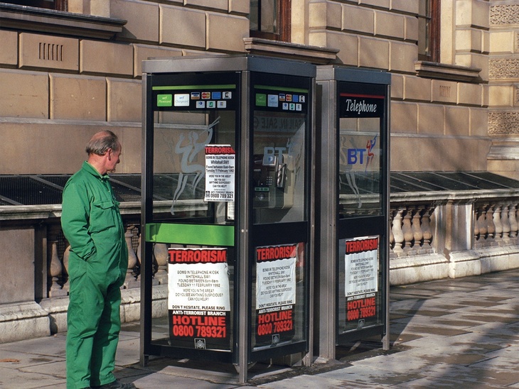 A man in green overalls surveys phone booths with terrorism hotline ads pasted in the windows