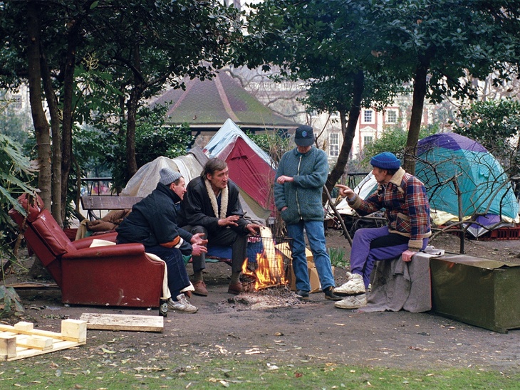 Man sit around a fire by tents in the middles of a park