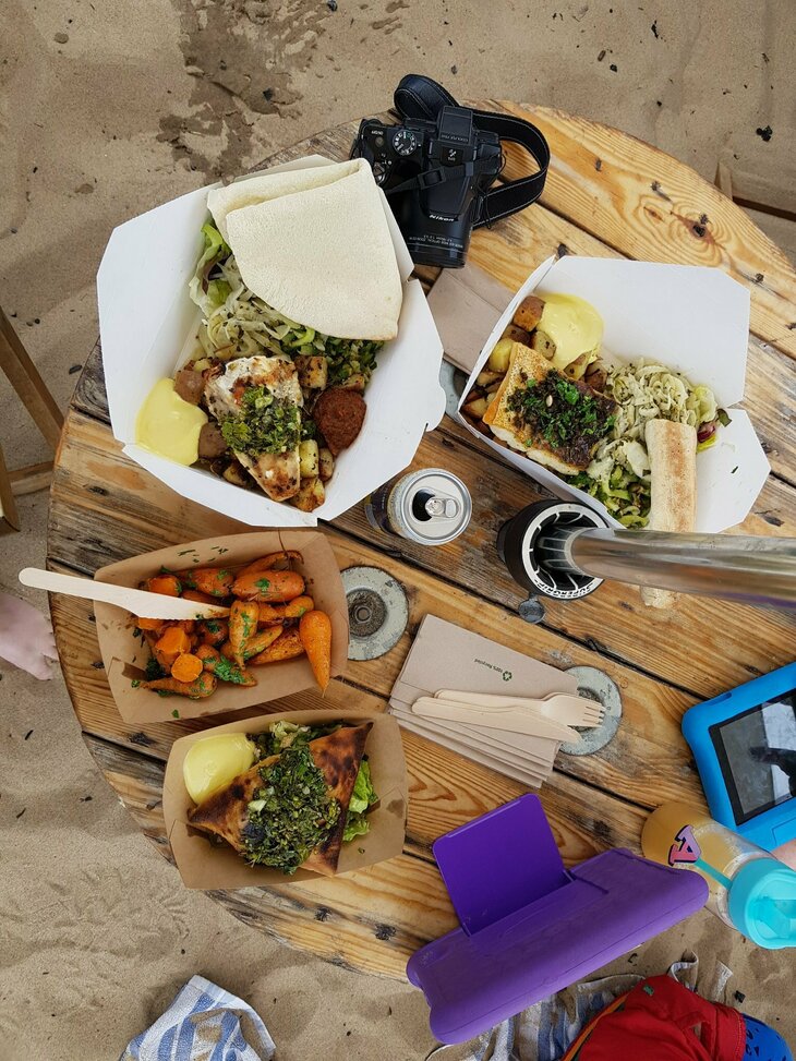 A selection of dishes filled with cooked fish, as well as a dish of carrots can be seen. The table is an old cable reel, and is placed on a sandy beach. 