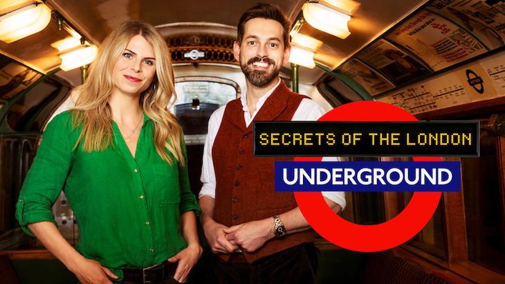 A female presenter in a green top and a male presenter in a red tank top stand inside a vintage tube carriage. A tube roundel superimposed in front says Secrets of the London Underground