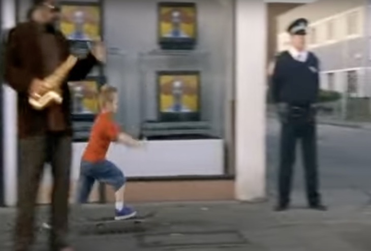 A boy in an orange shirt skateboards through a high street. A policeman watches from a corner